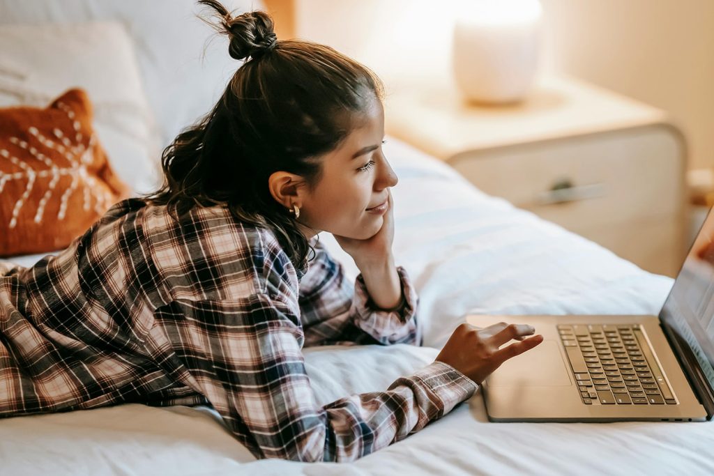Younger woman lays on bed reading on her laptop